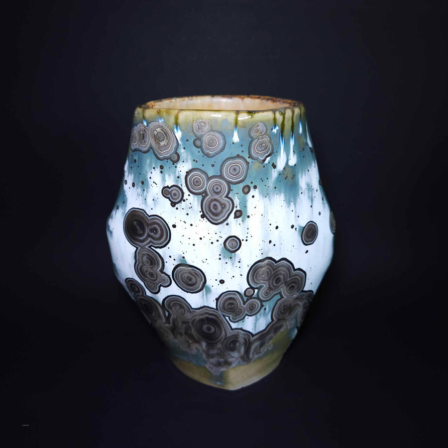 An octagonal vase with turquoise crystalline glaze. The crystals formed black and white concentric circles and are mostly clustered. Some appear as though they are dividing as in cell mitosis. The interior is glazed light yellow.