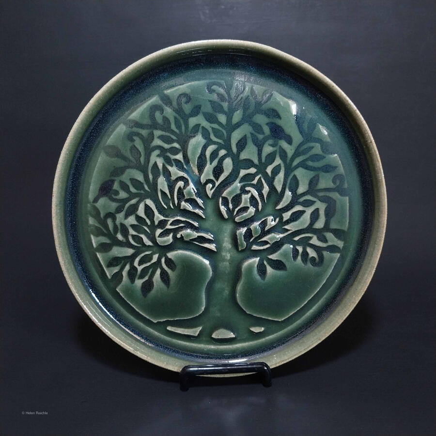 A round, green stoneware plate with a large tree impressed in center. The canopy and trunk extends to the edge of the plate and 2 little birds are amongst the leaves and branches.