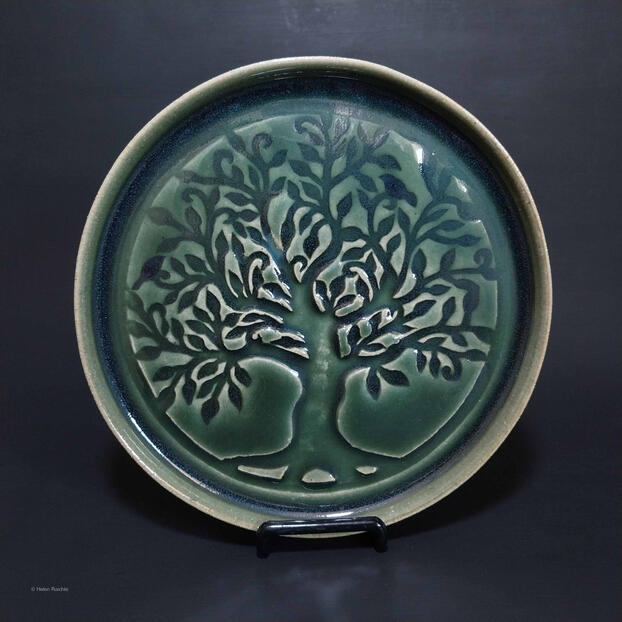 A round, green stoneware plate with a large tree impressed in center. The canopy and trunk extends to the edge of the plate and 2 little birds are amongst the leaves and branches.