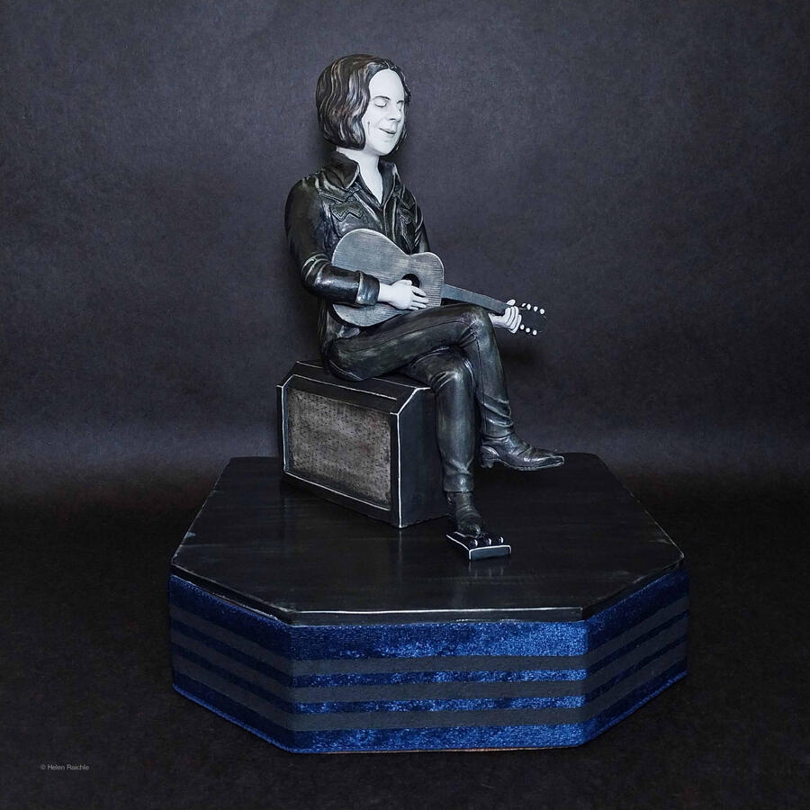 A musician sits cross-legged atop a rectangular amplifier. He holds an acoustic guitar with both hands and has closed eyes with a smile on his face. Under his foot is a sound effects pedal. The figure and his instruments rest atop a hexagonal shaped stage