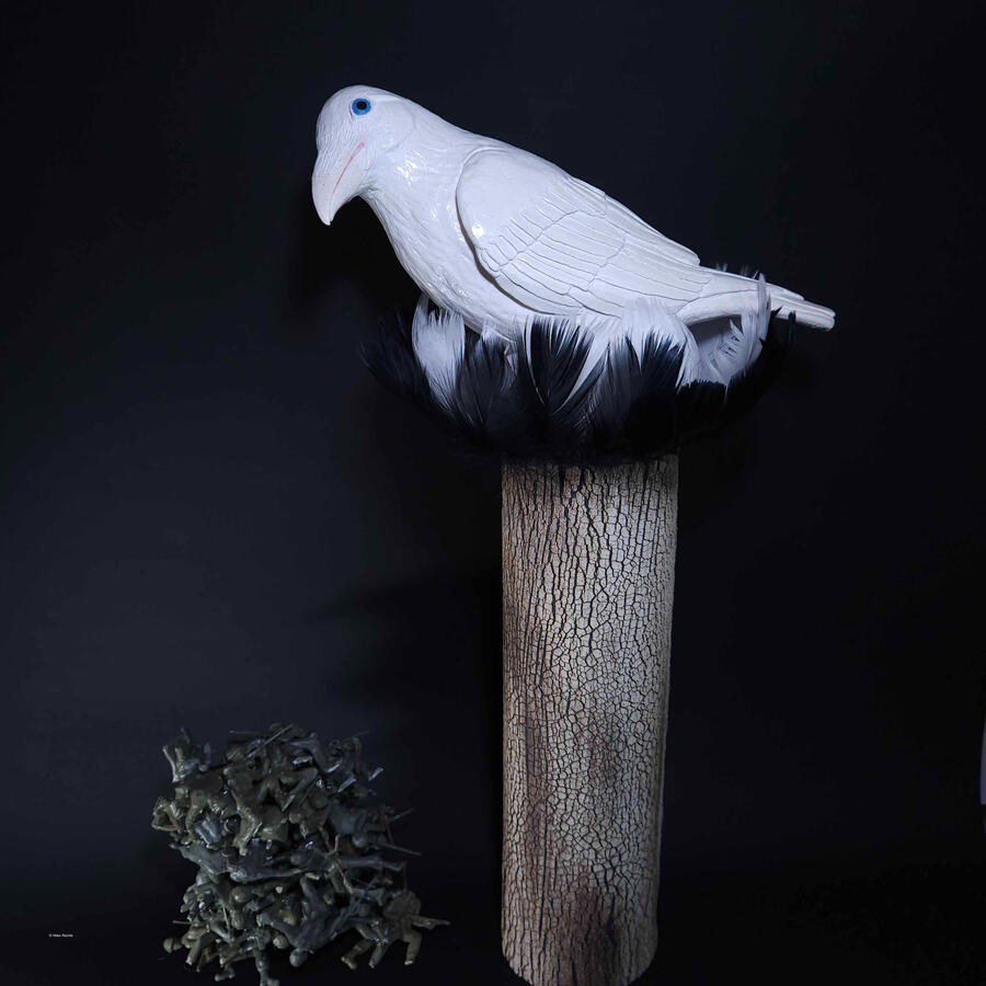 A white bird with blue eyes perched atop a tree bark textured cylinder. Real black and white feathers encircle the top of the cylinder and the legs of the bird. Below, 50 plastic army men lay in a pile.