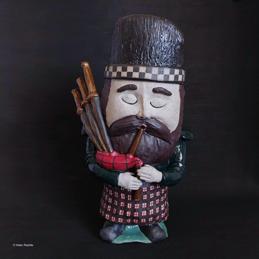A bearded, Scottish bagpiper garden gnome with a red, black, white plaid kilt, dark green shirt, and black and white checkered bonnet inflates his black and red plaid bagpipes