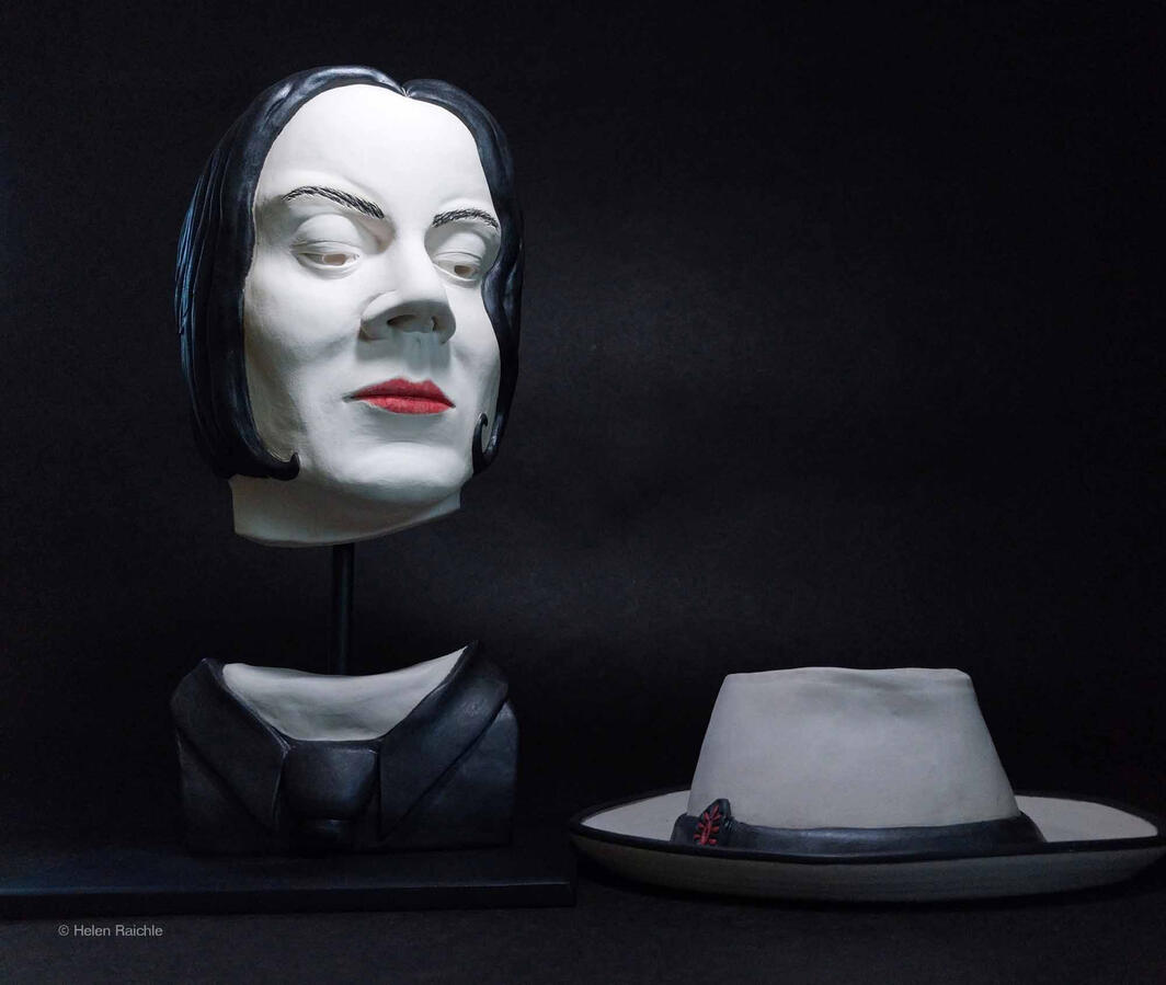 A bust of a man wearing a black suit and tie includes his head as a mask and the front of his chest. There is a gap where his neck should be. The bust is held up with a black steel rod and square base. The man gazes down to his fedora lying on the table.