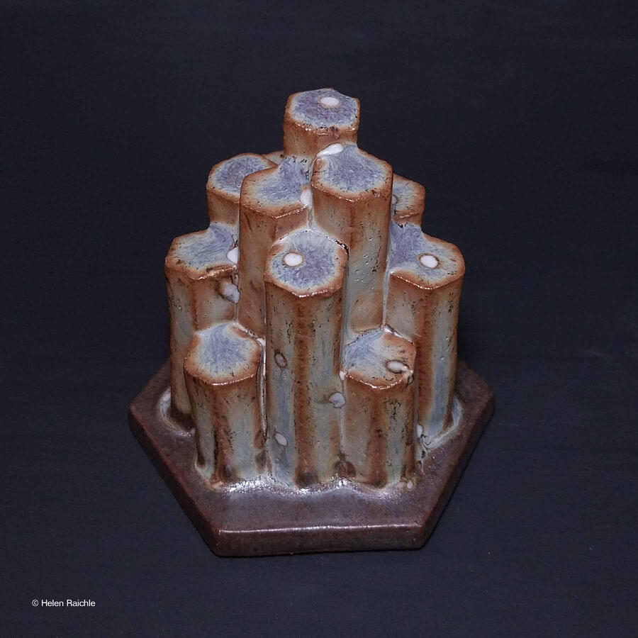 A group of hexagonal columns of different heights rise out of a hexagonal shaped base. The columns are blue-gray glazed with brown edges. There are white spots resembling lichen that grows on the rocks.