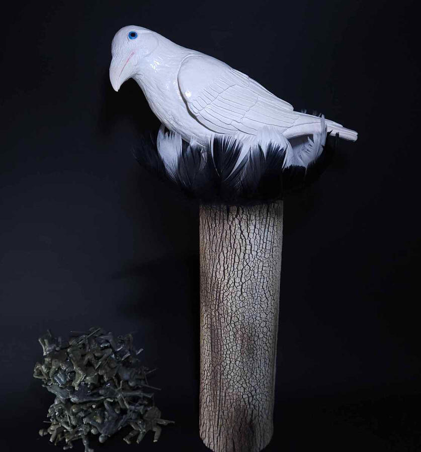 A white bird with blue eyes perched atop a tree bark textured cylinder. Real black and white feathers encircle the top of the cylinder and the legs of the bird. Below, 50 plastic army men lay in a pile.